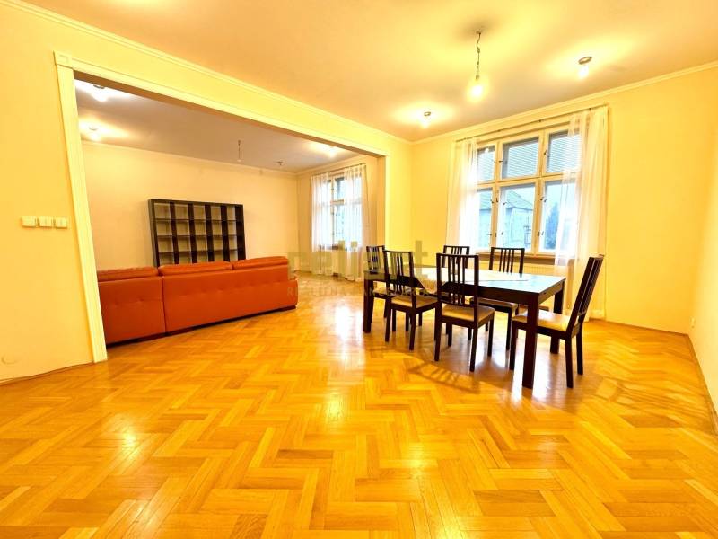 Jídelna se soupravou stolů a židlí, vedle obývacího pokoje s oranžovou sedačkou. Podlaha s dřevěným dekorem.