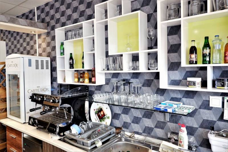 Interiér obchodních prostor s kávovarem, regály, sklenicemi a nápoji na poličkách.