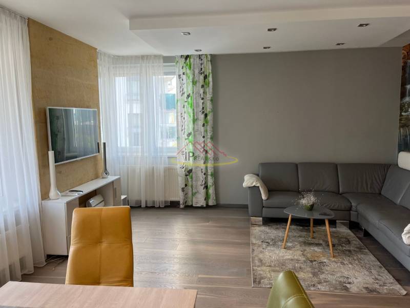 Obývací pokoj ve 4pokojovém bytě s podlahou s dřevěným dekorem, šedou sedačkou a TV.