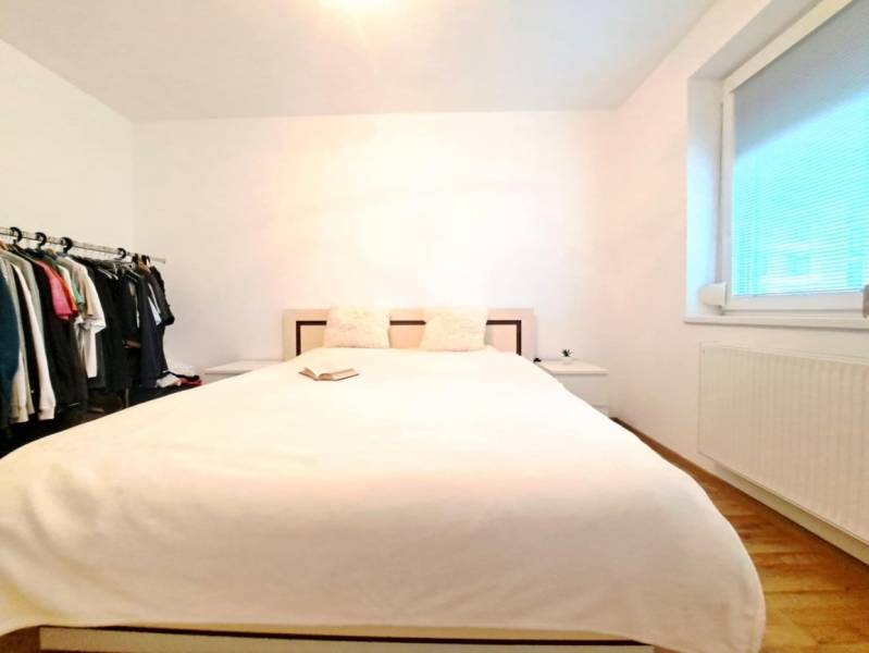 Ložnice ve dvoupokojovém bytě s postelí, otevřenou šatní skříní a podlahou s dřevěným dekorem.