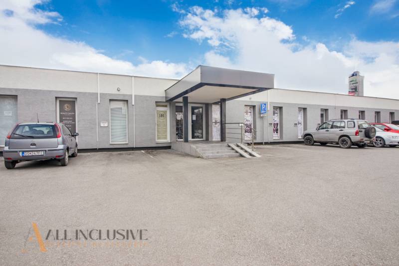 Jednopodlažní budova s obchodními prostory na ulici Vajanského, parkovištěm a auty před vstupem.