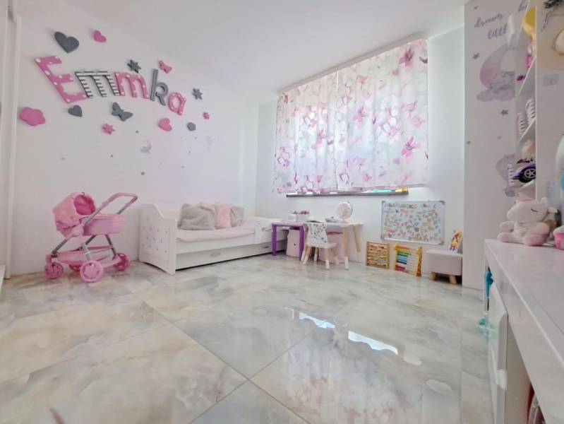 Dětský pokoj v rodinném domě s dekoracemi, kočárkem a psacími stoly. Podlaha má dřevěný dekor.