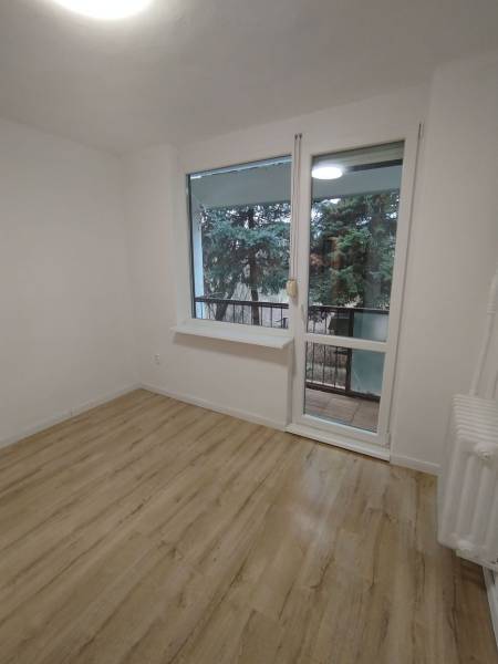 Pokoj se světlými stěnami, podlahou s dřevěným dekorem a výhledem na zeleň.