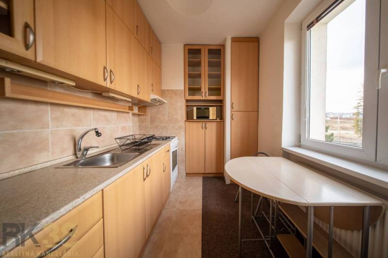 Kuchyně v 2-pokojovém bytě s podlahou s dřevěným dekorem, světlým nábytkem a stolem u okna.