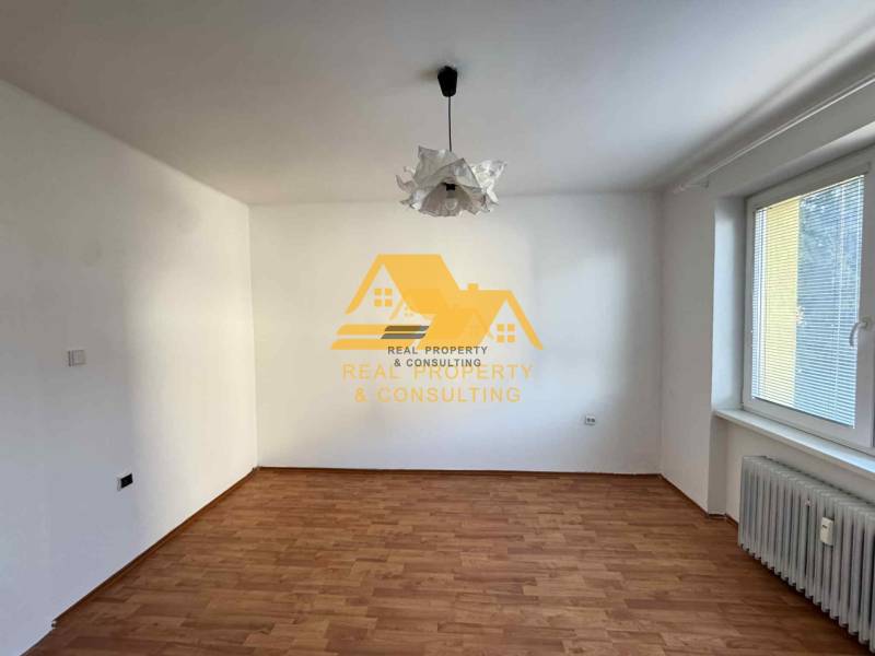 Pokoj v 3-pokojovém bytě s bílými stěnami, podlahou s dřevěným dekorem a radiátorem.