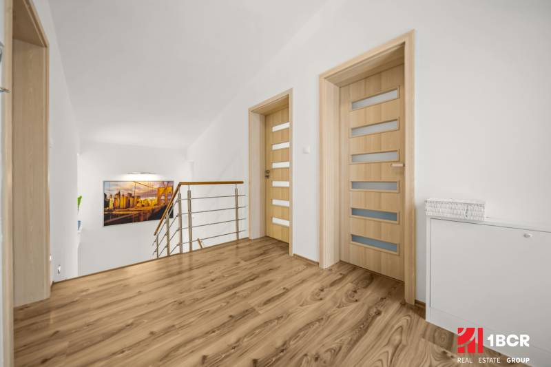 Chodba v rodinném domě s podlahou s dřevěným dekorem, minimalistickým zábradlím a uměleckou fotografií.