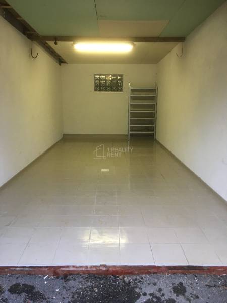 Pohled do prázdné garáže se šedou dlažbou na podlaze a regálem vzadu.