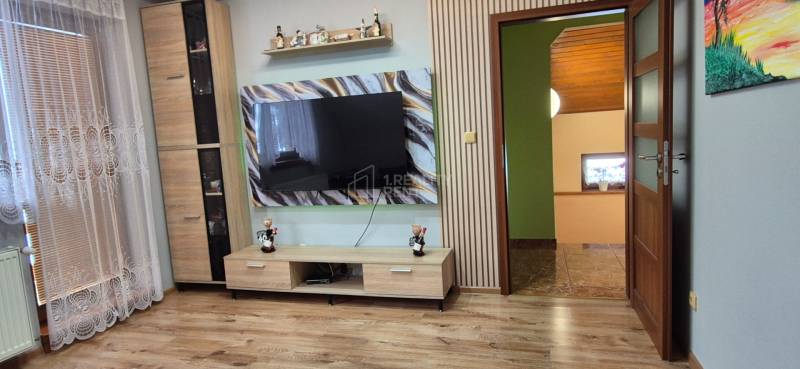 Obývací pokoj v rodinném domě s dřevěným dekorem podlahy a velkou televizí na stěně.