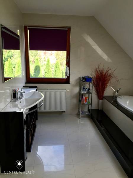 Koupelna v rodinném domě s vanou, umyvadlem, regálem a oknem s fialovou roletou.