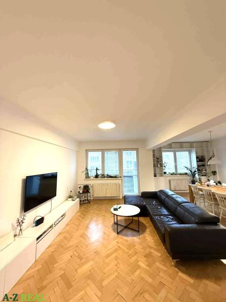 Obývací pokoj ve 4pokojovém bytě s podlahou s dřevěným dekorem a černou sedačkou.