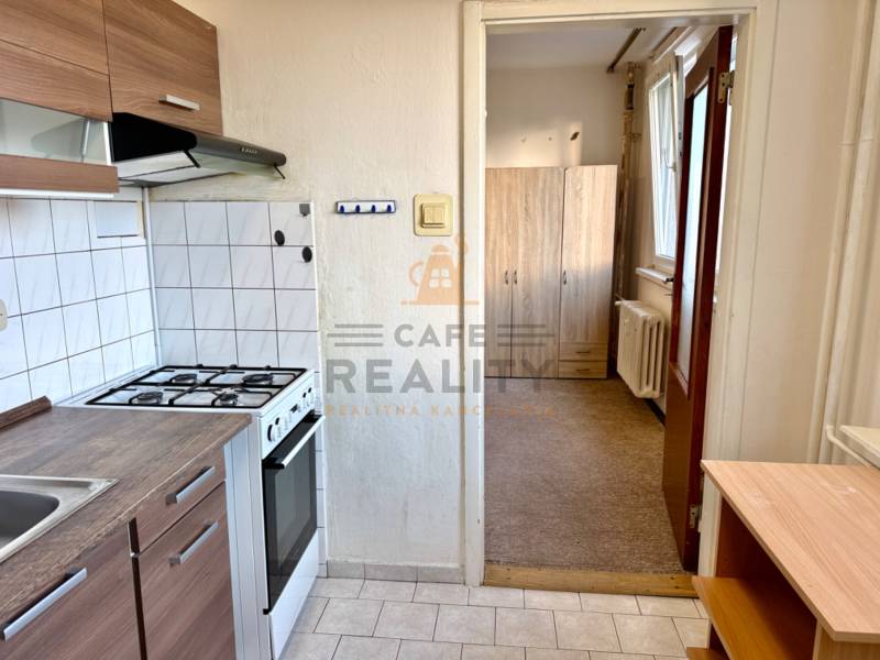 Kuchyně s bílými obkladačkami, sporákem, skříňkami a pohled do předsíně v 1-pokojovém bytě.