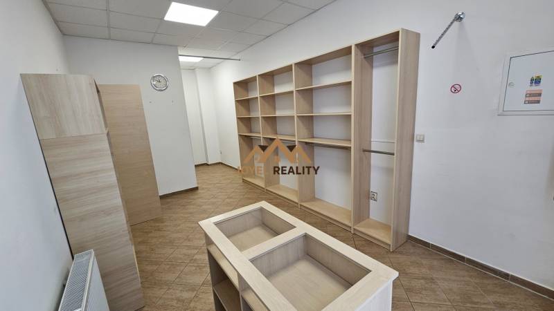 Obchodní prostory s regály a podlahou s dřevěným dekorem, bílá stěna, stropní osvětlení.