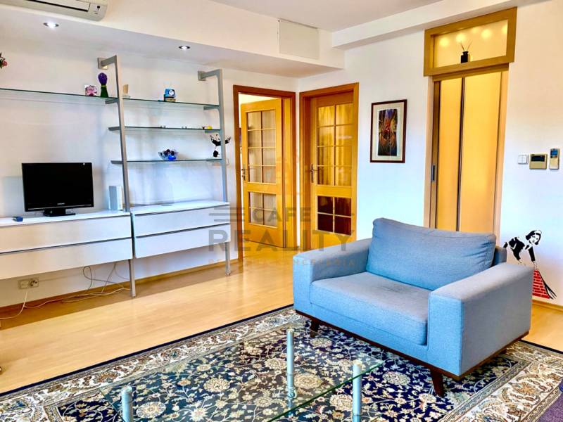 Obývací pokoj s pohovkou, poličkami, kobercem a podlahou s dřevěným dekorem ve 2pokojovém bytě.