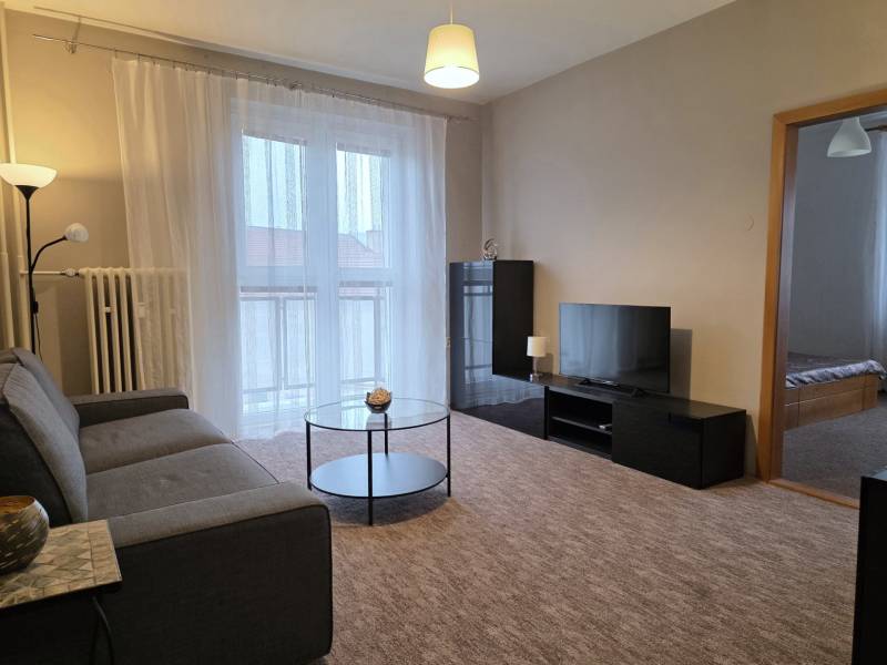 Obývací pokoj v 3-pokojovém bytě s pohovkou, televizí, stolkem a balkónovými dveřmi.