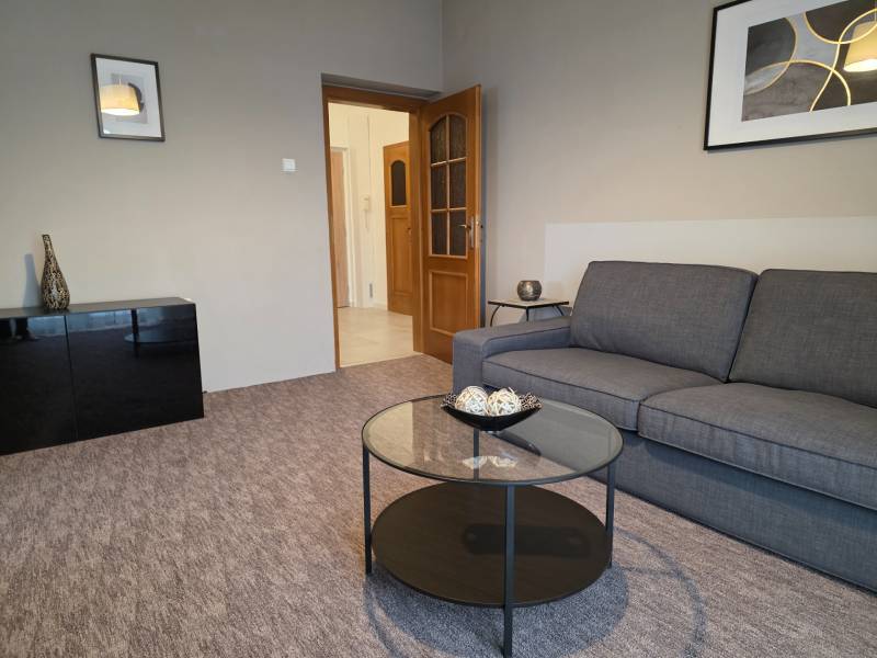 Obývací pokoj ve 3-pokojovém bytě s šedou sedačkou a proskleným stolem.