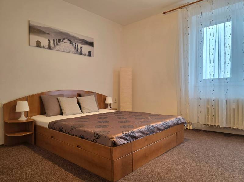 Ložnice v třípokojovém bytě s postelí, obrazem, nočními lampami a závěsy.