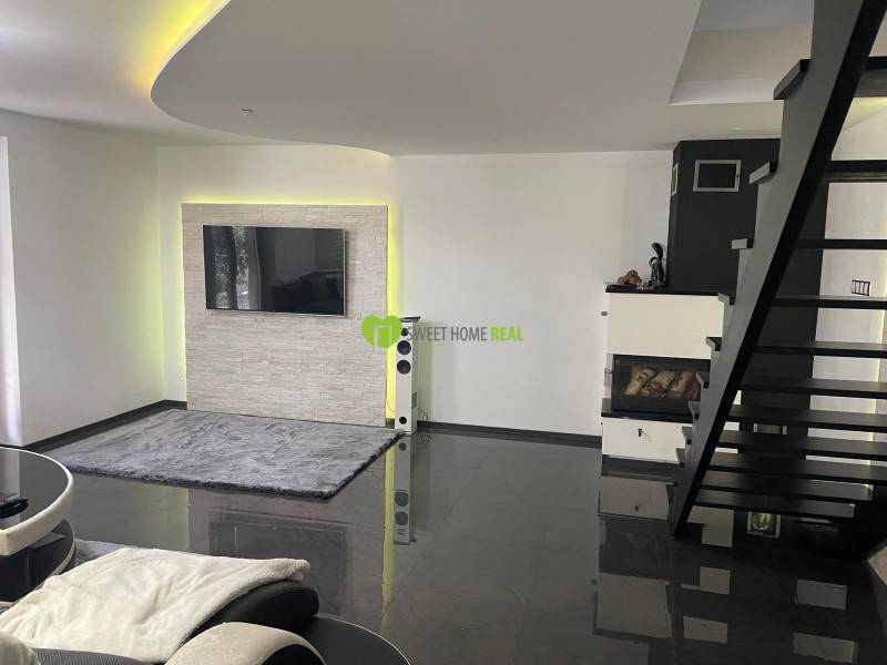 Obývací pokoj v rodinném domě se schodištěm, krbem a televizí na stěně.