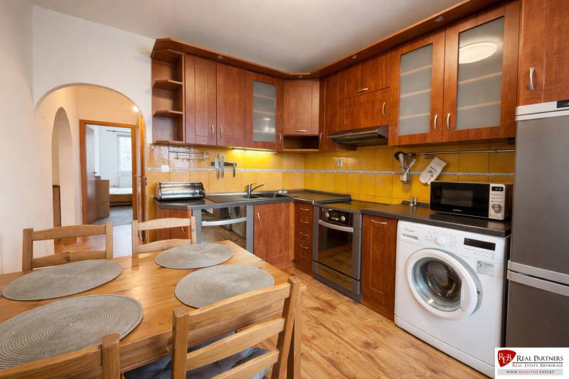 Kuchyně 4pokojového bytu s dřevěným dekorem, vybavená spotřebiči, jídelní stůl, žluté obklady.