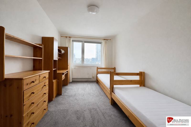 Světlý pokoj s dvěma postelemi, dřevěnými policemi a kobercem ve 4pokojovém bytě.