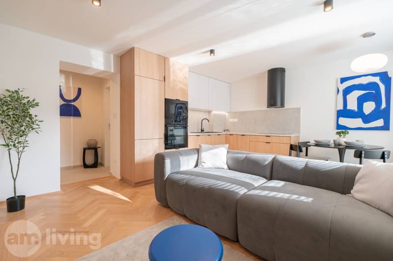 Obývací část 3pokojového bytu s kuchyní, šedou pohovkou a podlahou s dřevěným dekorem.