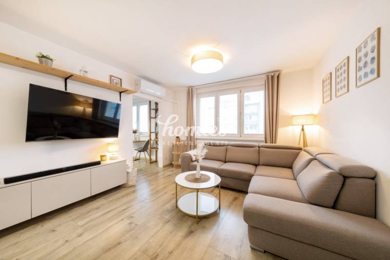 Obývací pokoj v 2pokojovém bytě s pohodlnou sedačkou a podlahou s dřevěným dekorem.