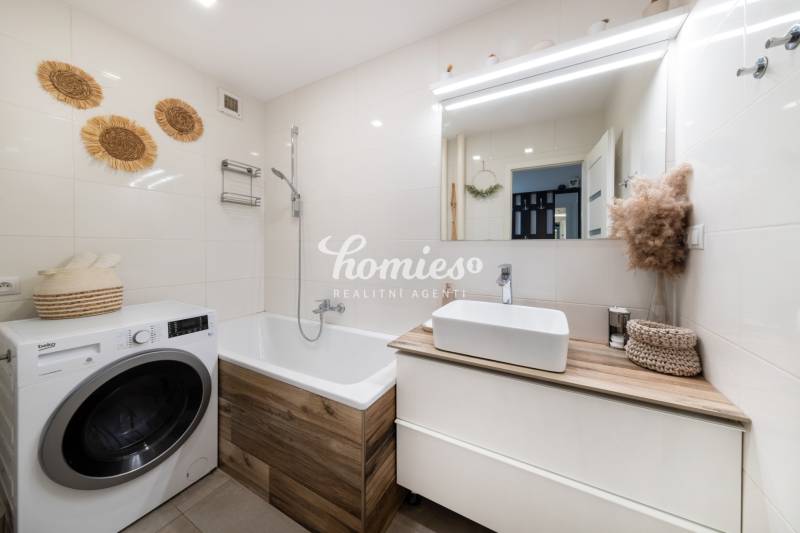 Koupelna v 2-pokojovém bytě s pračkou a vanou, světlé obklady a dekorace.