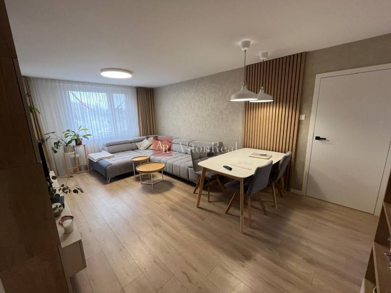 Obývací pokoj čtyřpokojového bytu s podlahou s dřevěným dekorem, šedou sedačkou a jídelním stolem.