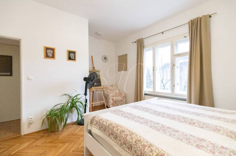 Světlá ložnice s podlahou s dřevěným dekorem, rostlinami a velkými okny ve 2pokojovém bytě.