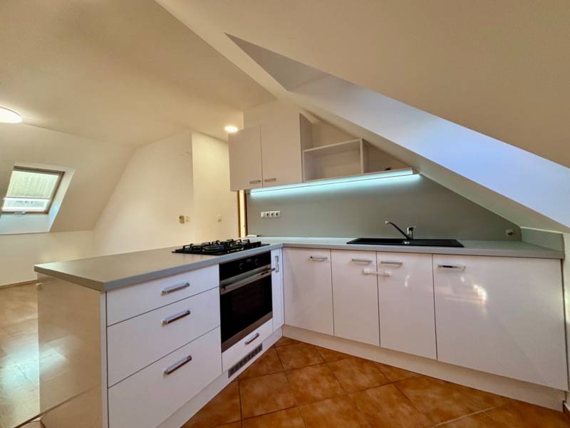 Světlá kuchyně s podkrovním oknem, rohovou pracovní deskou a keramickou dlažbou v 2-pokojovém bytě.