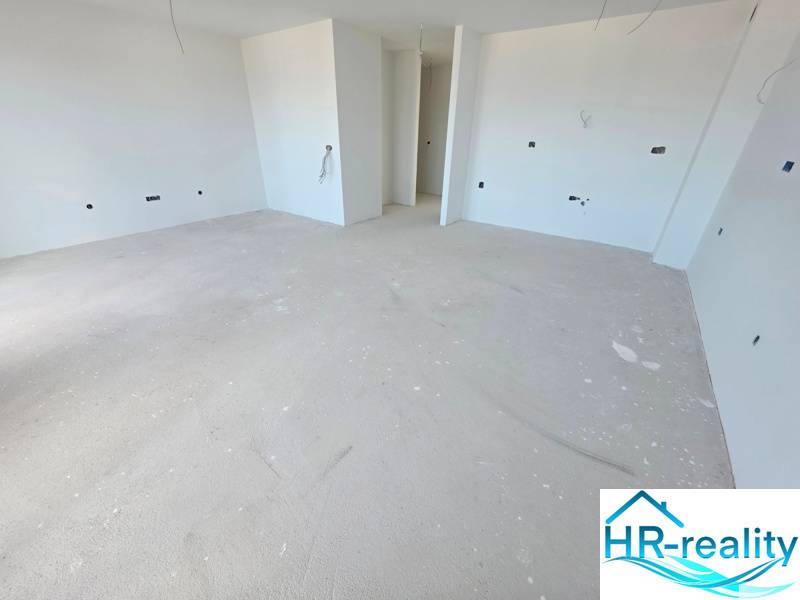 Prostorná nedokončená místnost v Rekreačním apartmánu s bílými stěnami a betonovou podlahou.