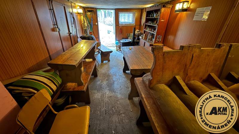 Interiér chaty s dřevěnými stoly a lavicemi, výhled na zahradu.