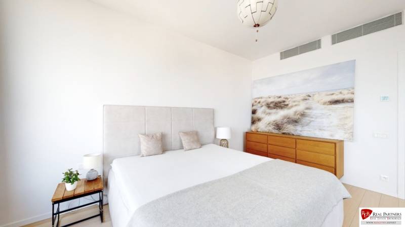 Ložnice v třípokojovém bytě s velkou postelí, obrazem a dřevěnou komodou.