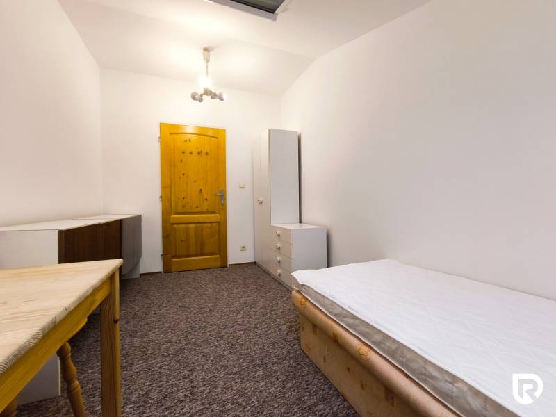 Pokoj v rodinném domě s postelí, skříní a stolem, doplněný bílým osvětlením a kobercovou podlahou.