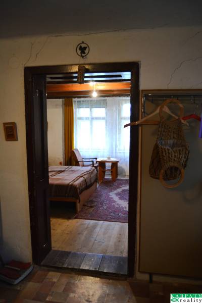 Interiér rodinného domu s postelí a dřevěnou podlahou, přes dveře je vidět pokoj.