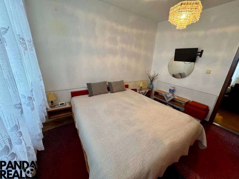 Ložnice v 3-pokojovém bytě s postelí, lustrem a televizorem na stěně.