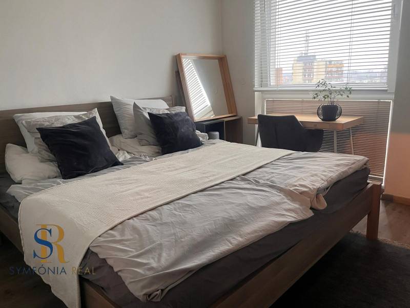 Ložnice v dvoupokojovém bytě s postelí, psacím stolem a podlahou s dřevěným dekorem.