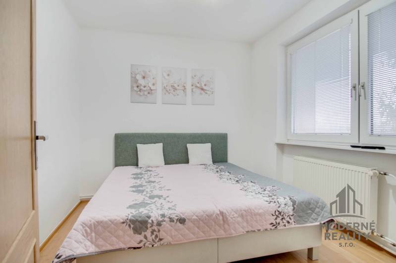 Ložnice se světlou postelí, podlahou s dřevěným dekorem a ozdobnými obrazy ve 4pokojovém bytě.