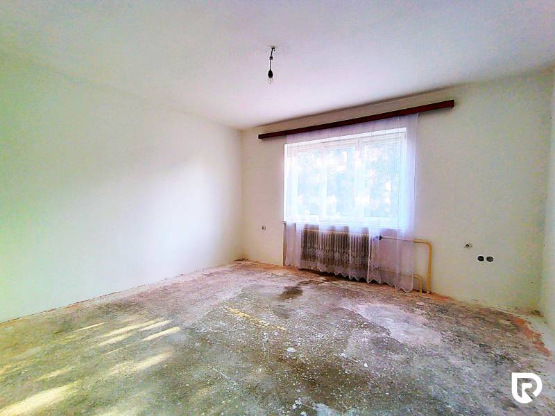 Prázdný pokoj v rodinném domě s opotřebovanou podlahou, radiátorem a závěsy.