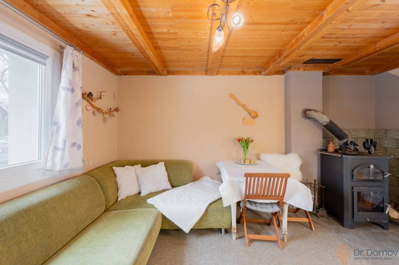 Interiér chaty s kachlovými kamny, zelenou sedačkou a stolem. Dřevěný strop a dekorace.