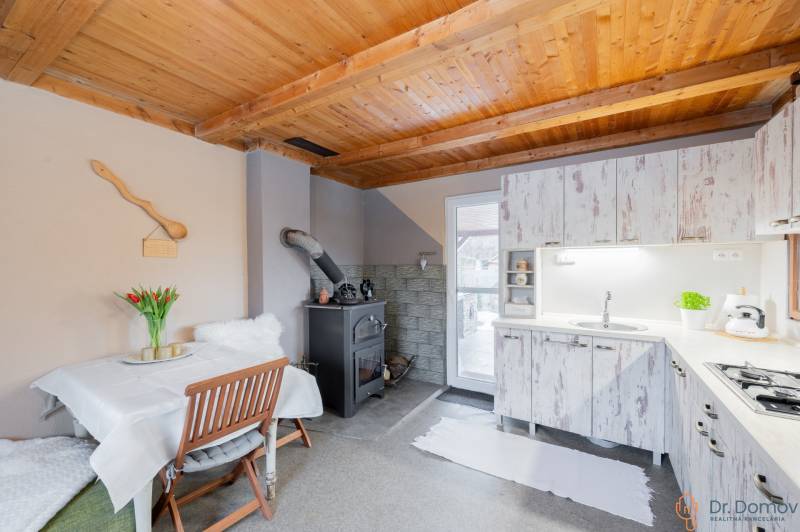Kuchyně v chatě s dřevěným stropem, pecí a bílým nábytkem.