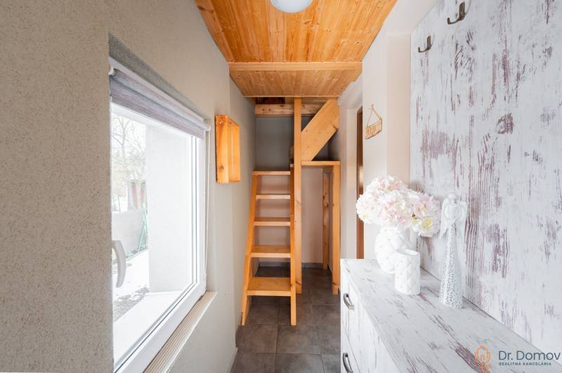 Interiér chaty s dřevěným schodištěm a oknem s výhledem na exteriér.