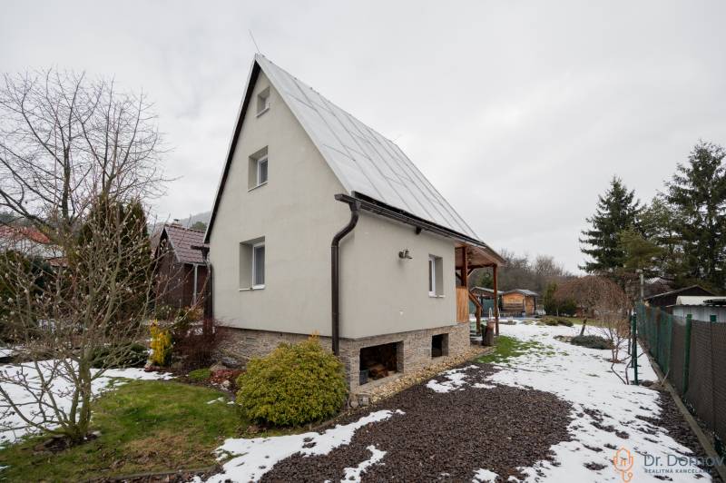 Chata v Handlové s trávníkem a zasněženou zahradou, obklopená stromy a keři.