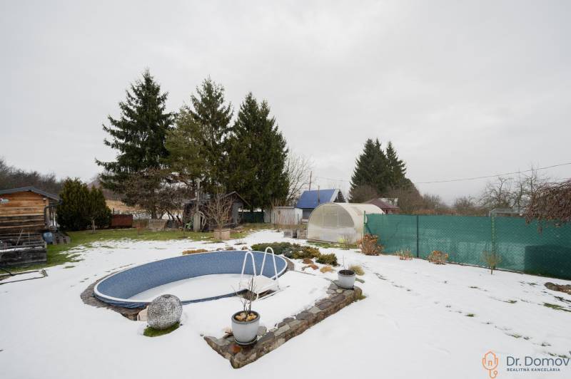 Zasněžená zahrada u chaty v Handlové s bazénem, skleníky a jehličnatými stromy.