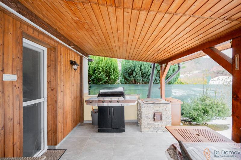 Dřevěné obložení a gril na terase chaty v Handlové, vedle zelených keřů.