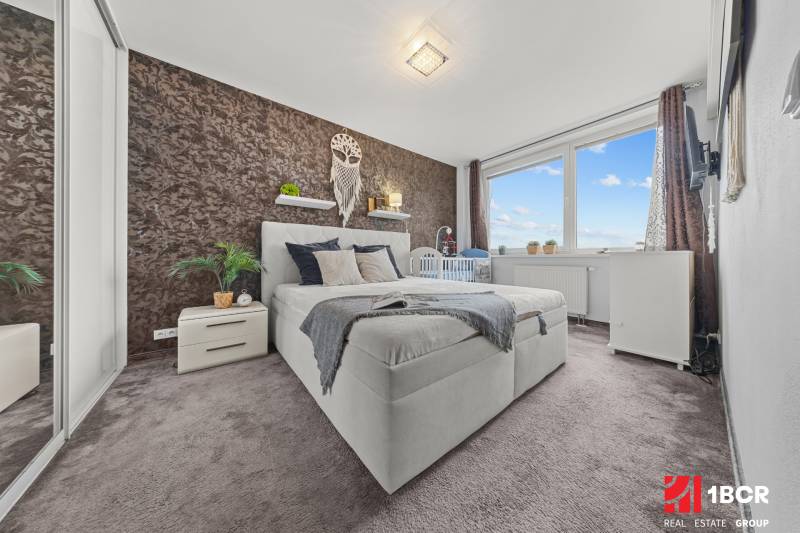 Ložnice v 2pokojovém bytě s manželskou postelí, kobercovou podlahou a dekorativní stěnou.