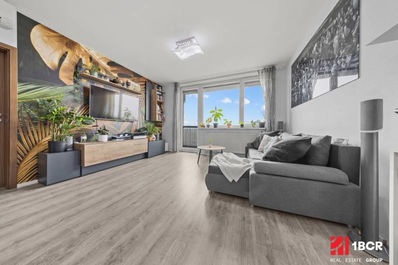 Obývací pokoj s pohodlným gaučem, tapetou s rostlinným motivem a podlahou s dřevěným dekorem.