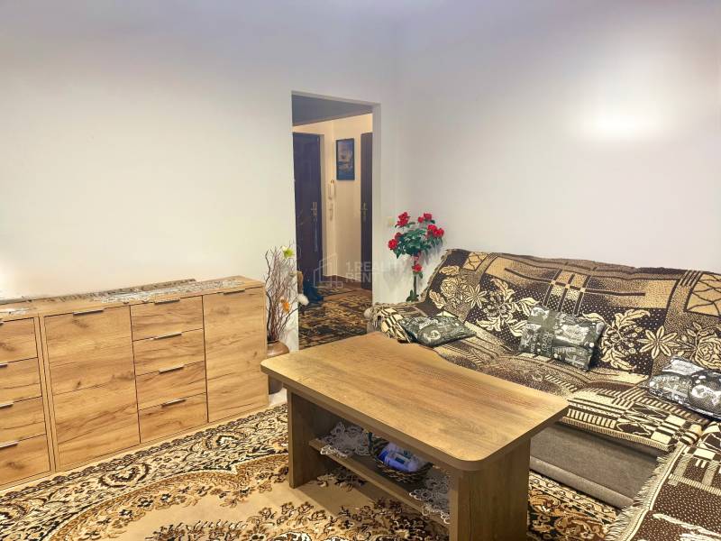 Obývací pokoj v dvoupokojovém bytě s dřevěnou komodou a vzorovaným kobercem.