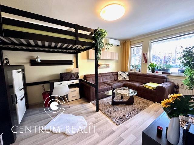 Obývací pokoj s patrovou postelí, sedačkou a podlahou s dřevěným dekorem v 1pokojovém bytě.