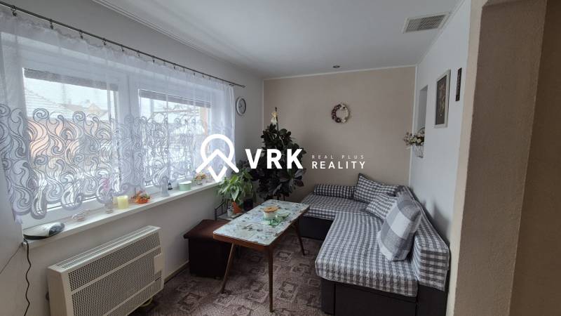 Obývací pokoj v rodinném domě s gaučem, stolem a oknem se záclonou.