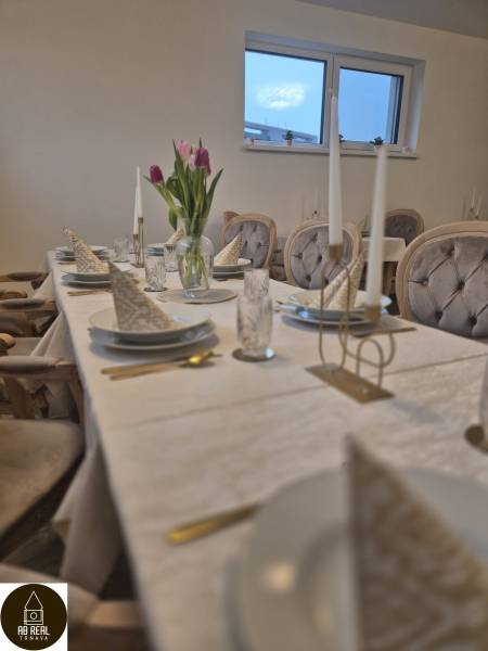 Stůl s bílým ubrusem, talíře, sklenice, květiny a potahy židlí v gastro prostorech.